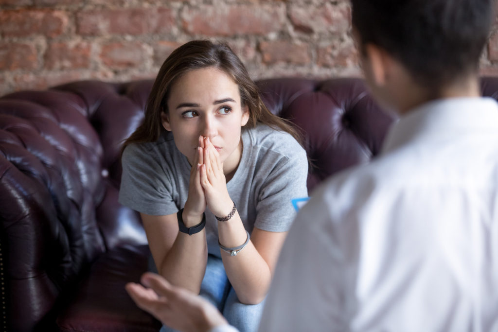 A young woman sitting on a couch, looking contemplative with her hands clasped together, while a counselor in a white shirt speaks to her, in a setting with a brick wall.